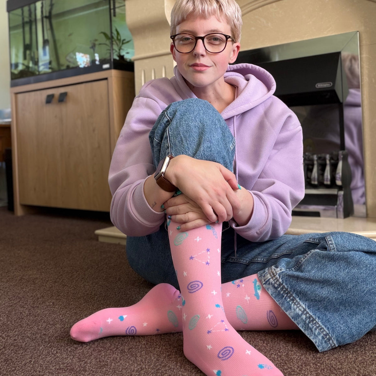 A person sitting on a carpeted floor in front of a fireplace, wearing pink knee-high socks with a colorful space-themed pattern of stars, planets, and spirals, along with a lavender hoodie and denim shorts.