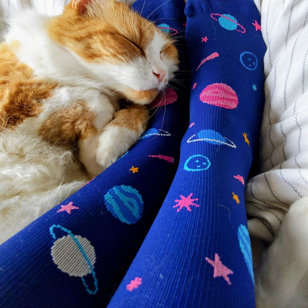 Cat laying on legs wearing navy blue knee high compression socks with space-themed design